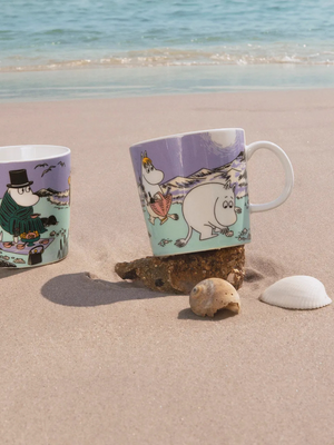 Two mugs with moomin characters on a sandy beach with ocean in the background